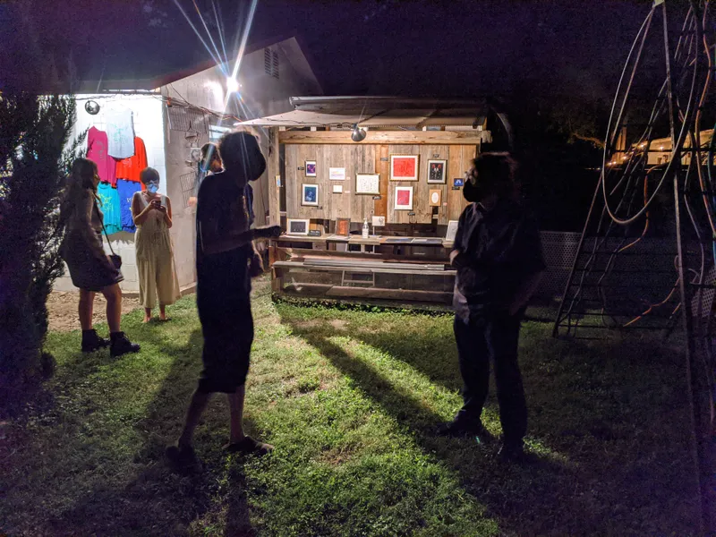 People gathered at an outdoor art exhibit at night, with displayed artwork and clothing for sale.