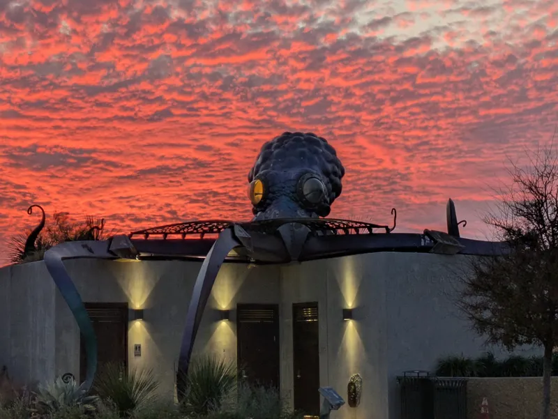 A large metal octopus sculpture sits atop a building with a vibrant red and orange sunset sky in the background.