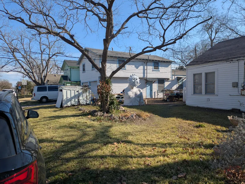 A large, white, sun-faced bust sculpture sits in a grassy yard in front of a white house.
