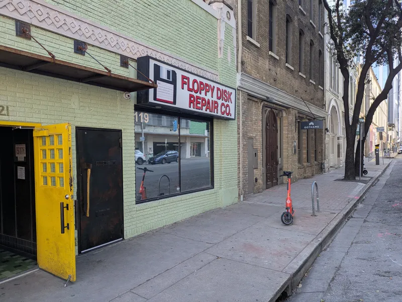 Floppy Disk Repair Co. storefront with a bright yellow door and a red electric scooter parked nearby.