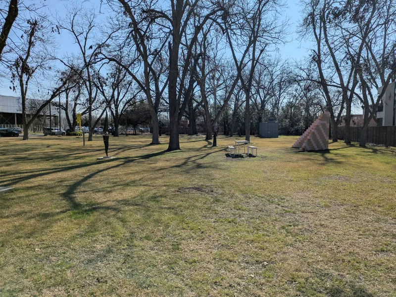 A park with bare trees, a picnic table, and a pink and yellow geometric sculpture under a clear blue sky.