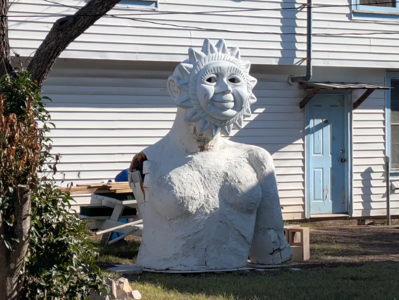 A large, white, unfinished statue of a sun-faced figure with a human-like torso stands outdoors in front of a white clapboard building.