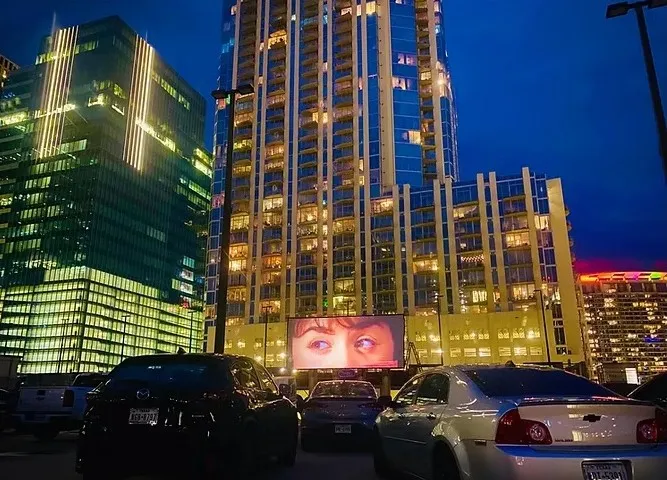 Cars parked in front of a drive-in movie screen showing a close-up of eyes, with tall buildings in the background at dusk.