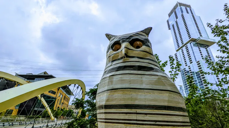 A large owl sculpture stands in front of a modern skyscraper and a curved bridge.