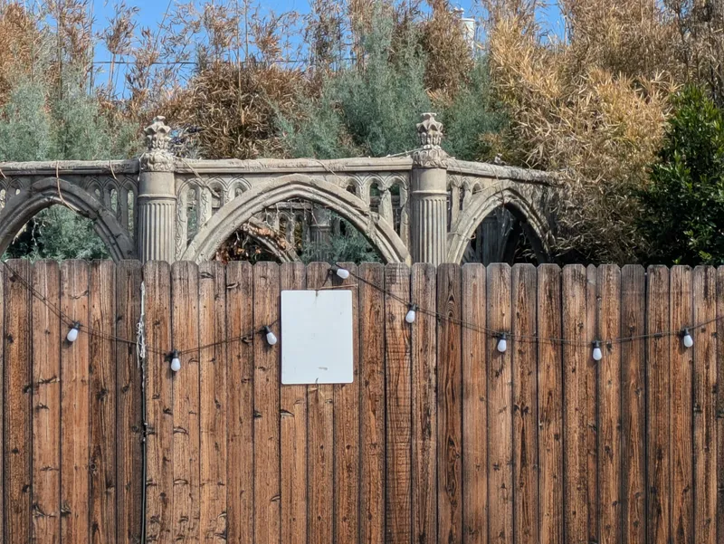 A rustic wooden fence with a blank white sign and string lights, with an ornate stone archway and foliage visible above.