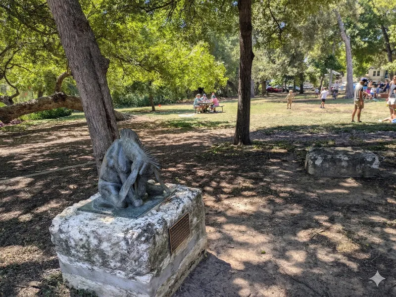 A sculpture of a creature sits on a stone base in a park, with people picnicking and playing in the background.