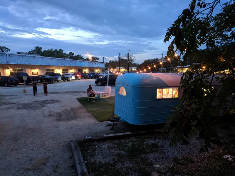 A blue vintage trailer with lights on sits in a gravel lot next to a building with cars parked outside.