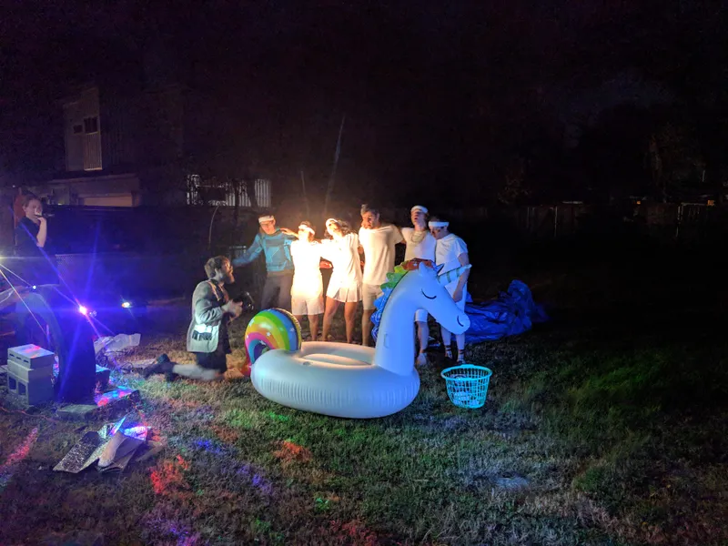 A group of young adults in white outfits and headbands pose with an inflatable unicorn at night.