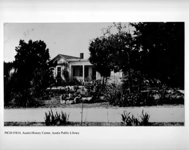 A black and white photo of a single-story house with a porch, surrounded by trees and a garden.