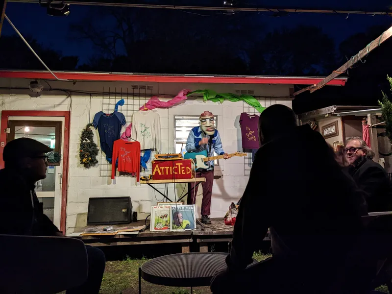 A performer in a mask plays guitar on a small stage at night, with t-shirts displayed on a wall behind.