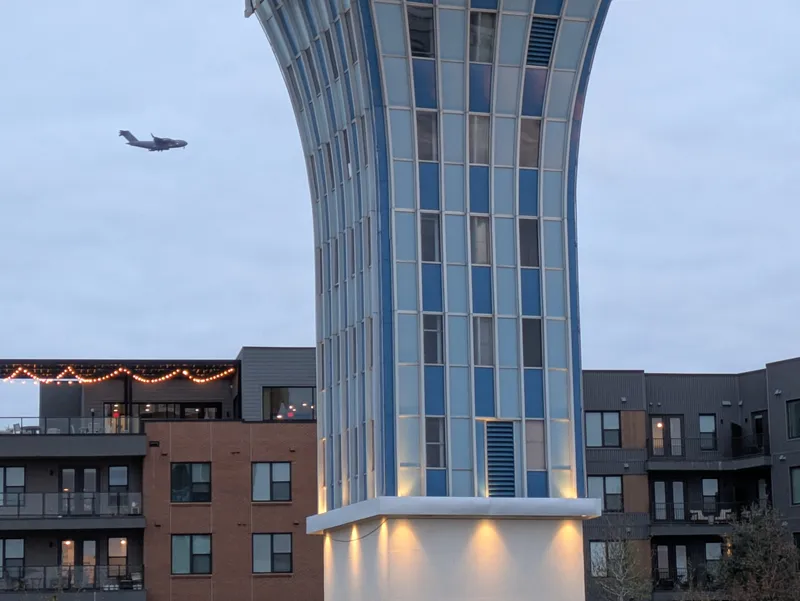 A large cargo plane flies in the sky near a modern building with blue and white windows.