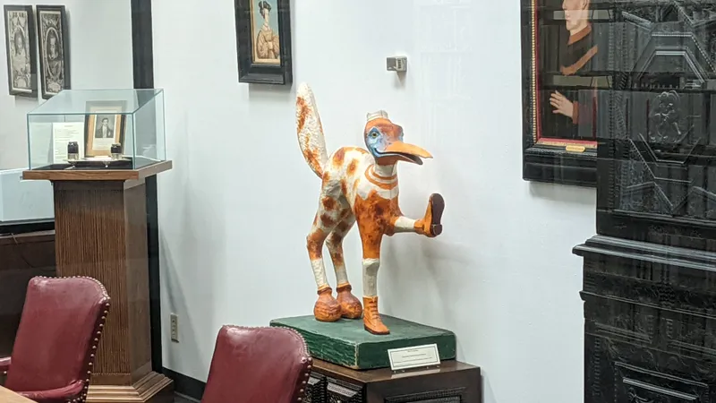 A whimsical sculpture of a bird-like creature with orange and white spots, wearing boots and a hat, in a museum-like setting.