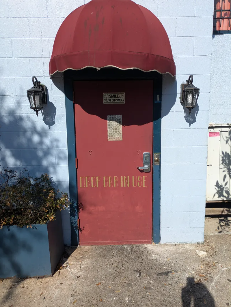 Red door with gold lettering "DROP BAR IN USE" under a burgundy awning, flanked by lanterns.