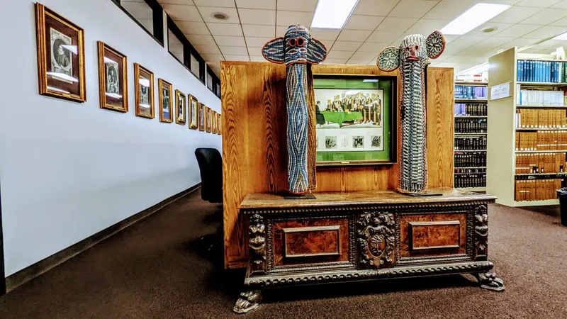 Two tall, beaded masks flank a framed document on an ornate wooden chest in a library. Framed portraits line a nearby wall.