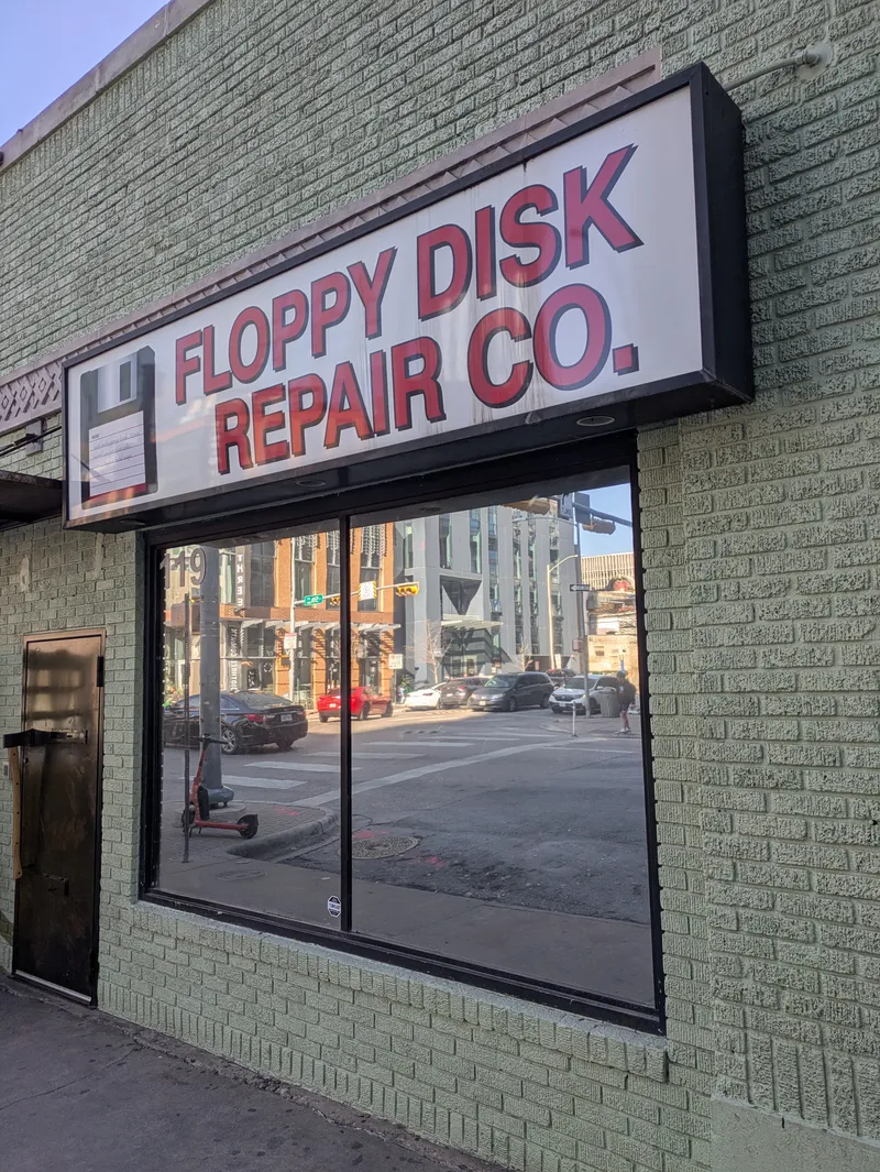 Floppy Disk Repair Co. sign on a light green brick building with a large window reflecting a city street.