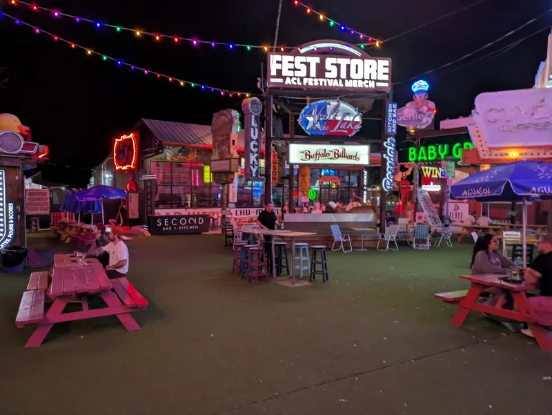 Outdoor seating area at night with colorful string lights and neon signs for various businesses.